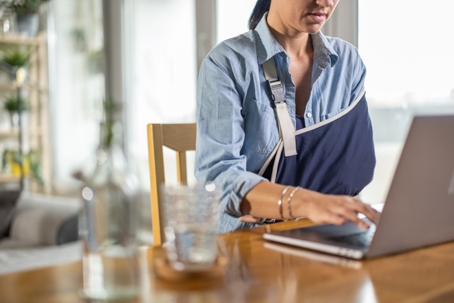 Unrecognizable woman with injured arm sitting at the table and using laptop at home.